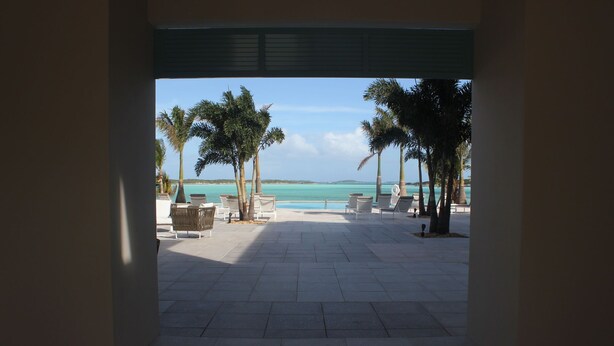 A view of a beachside lounge area with white lounge chairs and palm trees, leading to a pool and the ocean. - February Point Resort Estates BB1 (George Town)