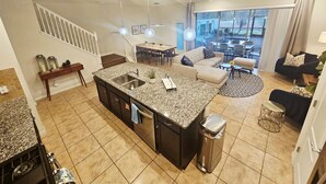 A modern kitchen with a central island, a dining area with a table and chairs, a living room with sofas, and a view through large windows. - 10% off Fall 2026 5-Bedroom Luxury Retreat w/ Modern Updates & Premium Amenities (Davenport)