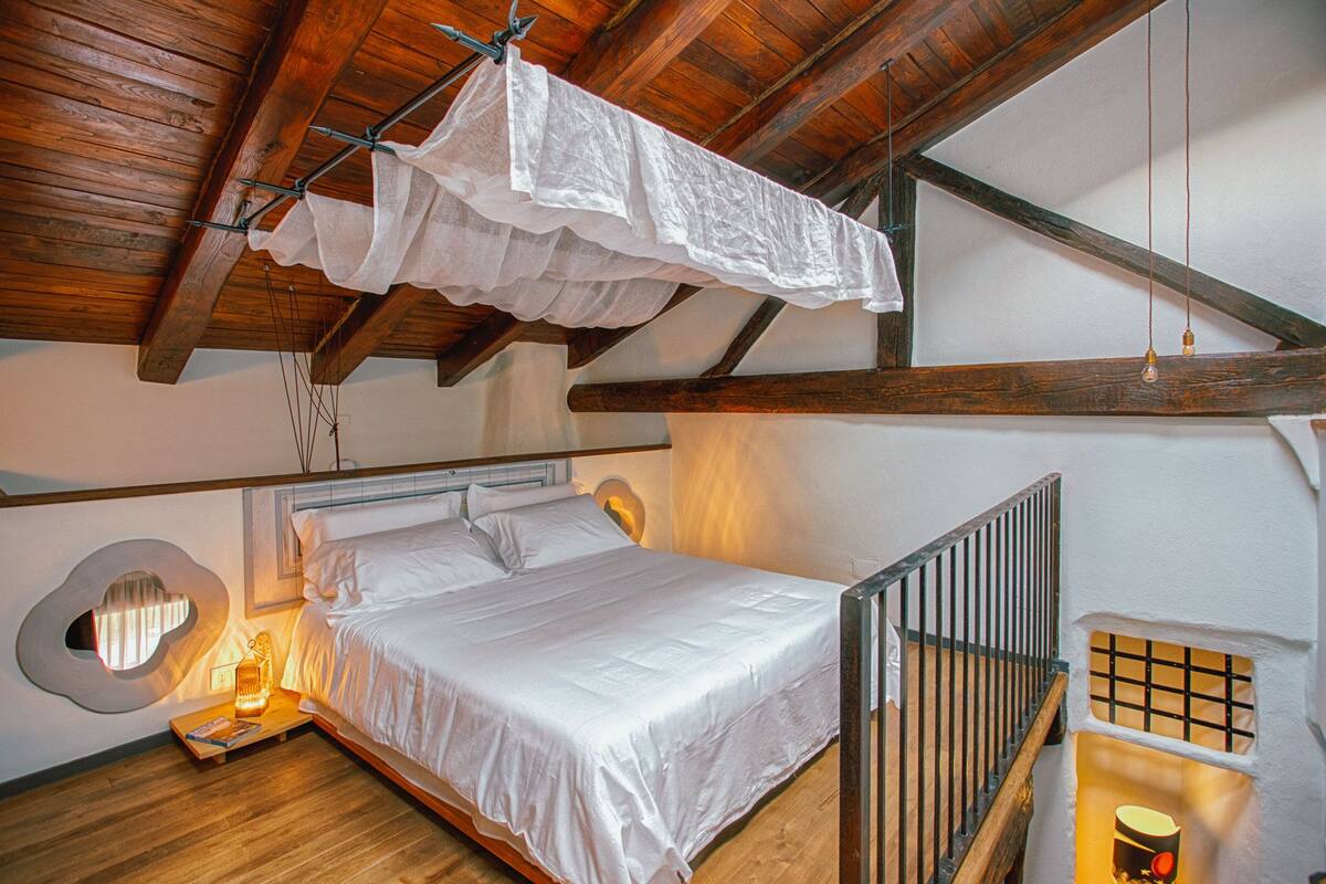 A bedroom with a wooden ceiling, a bed with white linens, a hanging laundry rack, a lantern, and a small table with a book.