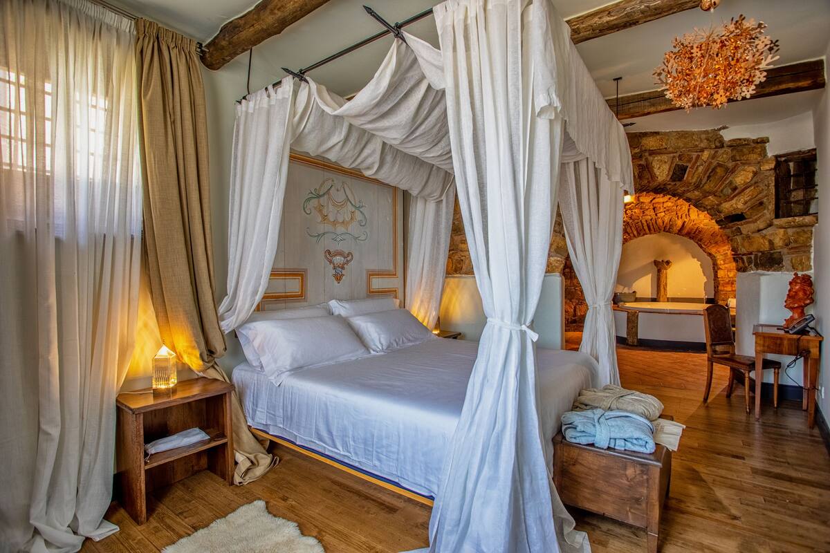 A bedroom with a canopy bed, wooden flooring, a stone fireplace, and a rustic stone wall.