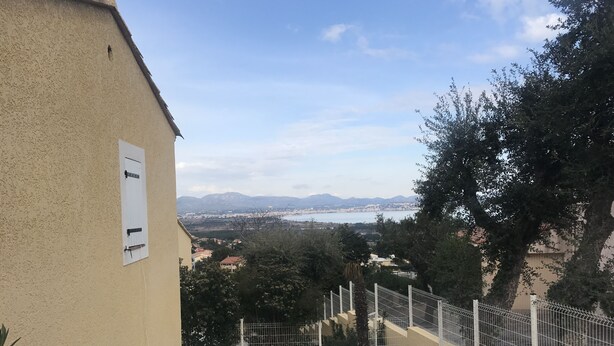 Vista desde un balcón con vista a la costa, cielo despejado, una ciudad lejana y un jardín cercado.