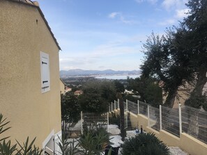 Vista desde un balcón con vista a la costa, cielo despejado, una ciudad lejana y un jardín cercado.