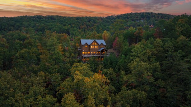 Une grande maison en bois à deux étages, entourée d’une forêt dense, avec une terrasse bien visible et de nombreuses fenêtres.