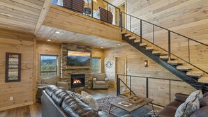 L’intérieur d’une cabane avec une cheminée en pierre, des canapés en cuir, un escalier et une grande fenêtre donnant sur un paysage montagneux.