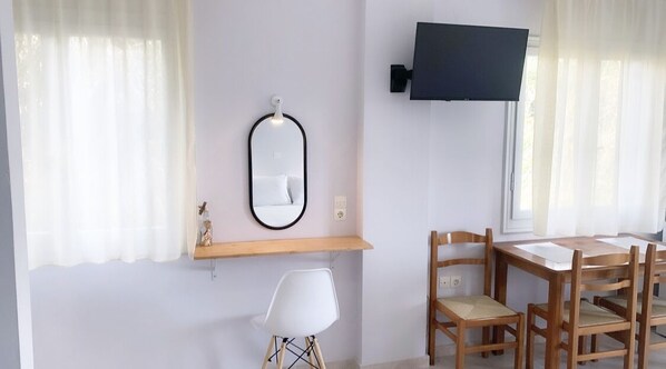 Una habitación con una mesa de comedor de madera, cuatro sillas, una silla blanca, un estante pequeño con un espejo y un televisor de pantalla plana montado en la pared.