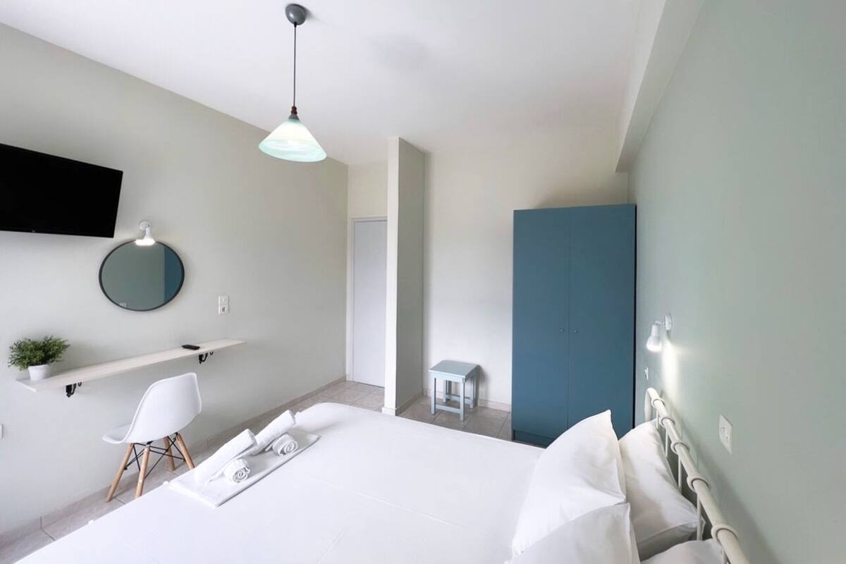 A modern hotel room with a large bed, a desk with a chair, a small table, a round mirror, and a TV mounted on the wall.
