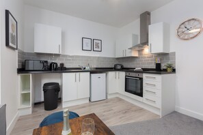 A modern kitchen with white cabinets, a black countertop, and a stainless steel range hood. There is a black trash bin and a small refrigerator. - Charnley House Apartment 3 (Blackpool)