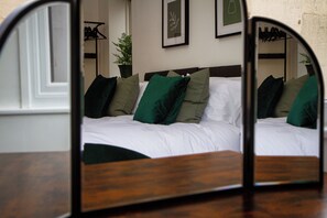 A bedroom with a wooden headboard, a bed with white bedding and green pillows, a potted plant, and a framed picture on the wall.