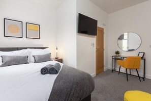 A modern hotel room with a neatly made bed, a desk with a chair, a television, and a round mirror on the wall.