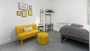 Una camera da letto moderna con un divano giallo, un letto grigio e un poggiapiedi rotondo.