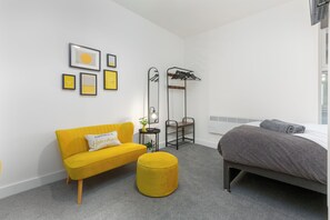 Un dormitorio moderno con un sofá amarillo, una cama gris y un otomano pequeño y redondo.