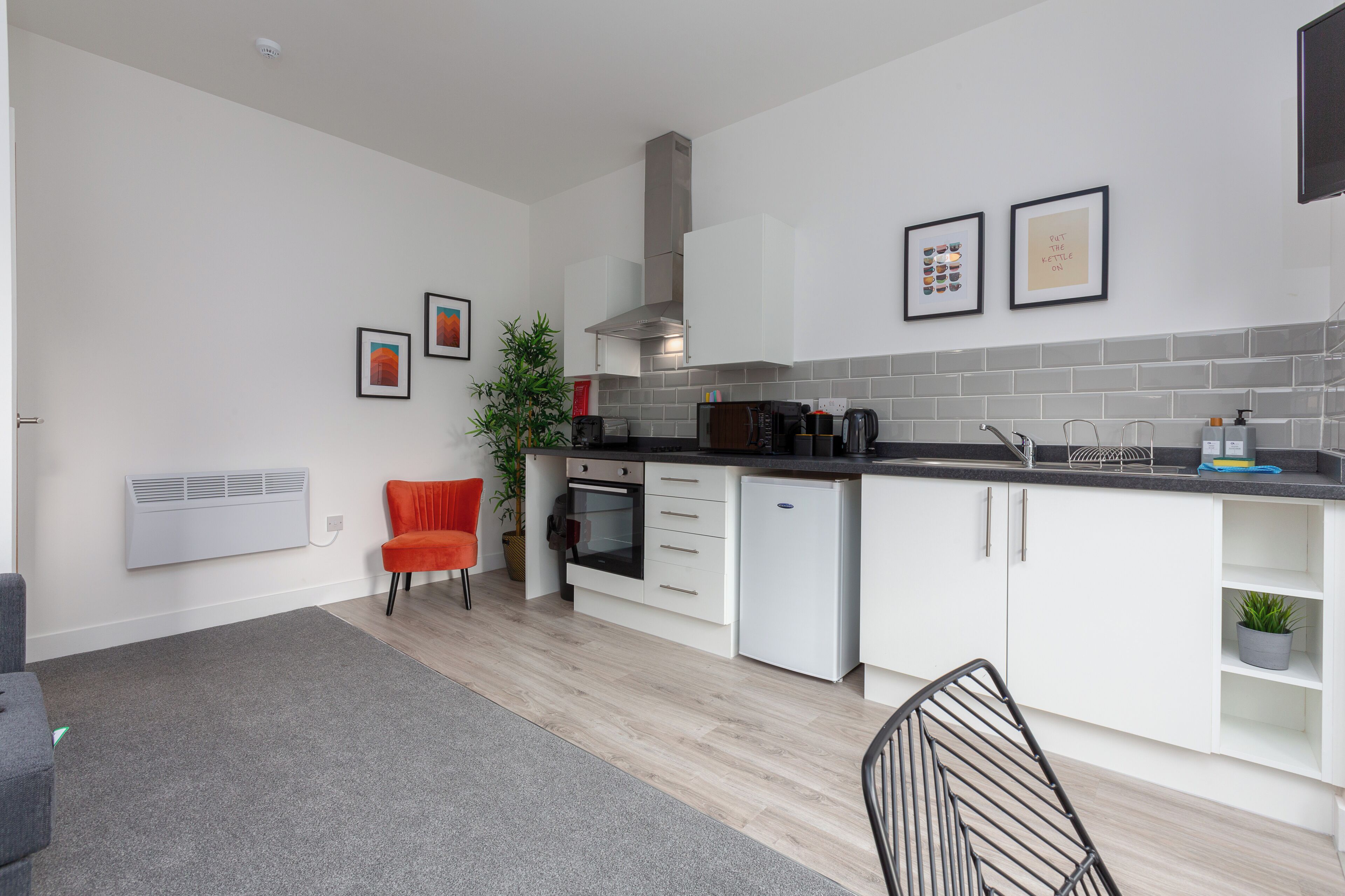 A modern kitchen with white cabinets, a grey tiled backsplash, and a black countertop. There are framed pictures on the wall, a potted plant, and a red chair.