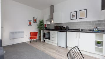 A modern kitchen with white cabinets, a grey tiled backsplash, and a black countertop. There are framed pictures on the wall, a potted plant, and a red chair.