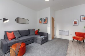 A modern living room with a grey sectional sofa, a red armchair, and a black floor lamp. There are framed artworks on the wall and a white radiator.