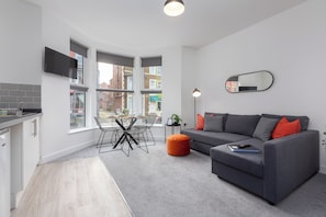 Eine moderne Wohnung mit Küche, Essbereich und Wohnzimmer. Sie verfügt über große Fenster, ein graues Sofa und einen Flachbildfernseher.