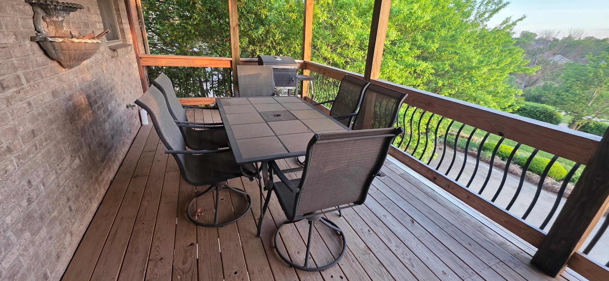 A wooden deck with a table and chairs, a grill, and a view of greenery.