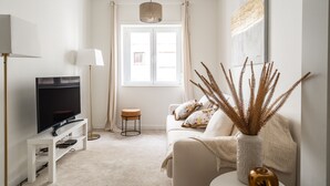 A modern living room with a flat-screen TV on a white stand, a sofa with cushions, a small wooden stool, and a decorative vase with dried plants. - Apartment (Lisboa)