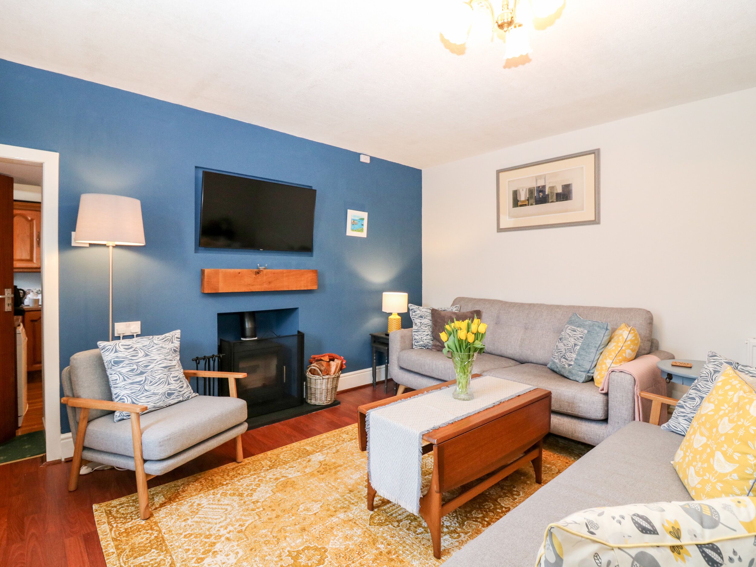 Una sala de estar con una pared azul de acento, un televisor de pantalla plana montado sobre una chimenea, un sofá gris con cojines estampados, una mesa de centro de madera y una lámpara de pie.