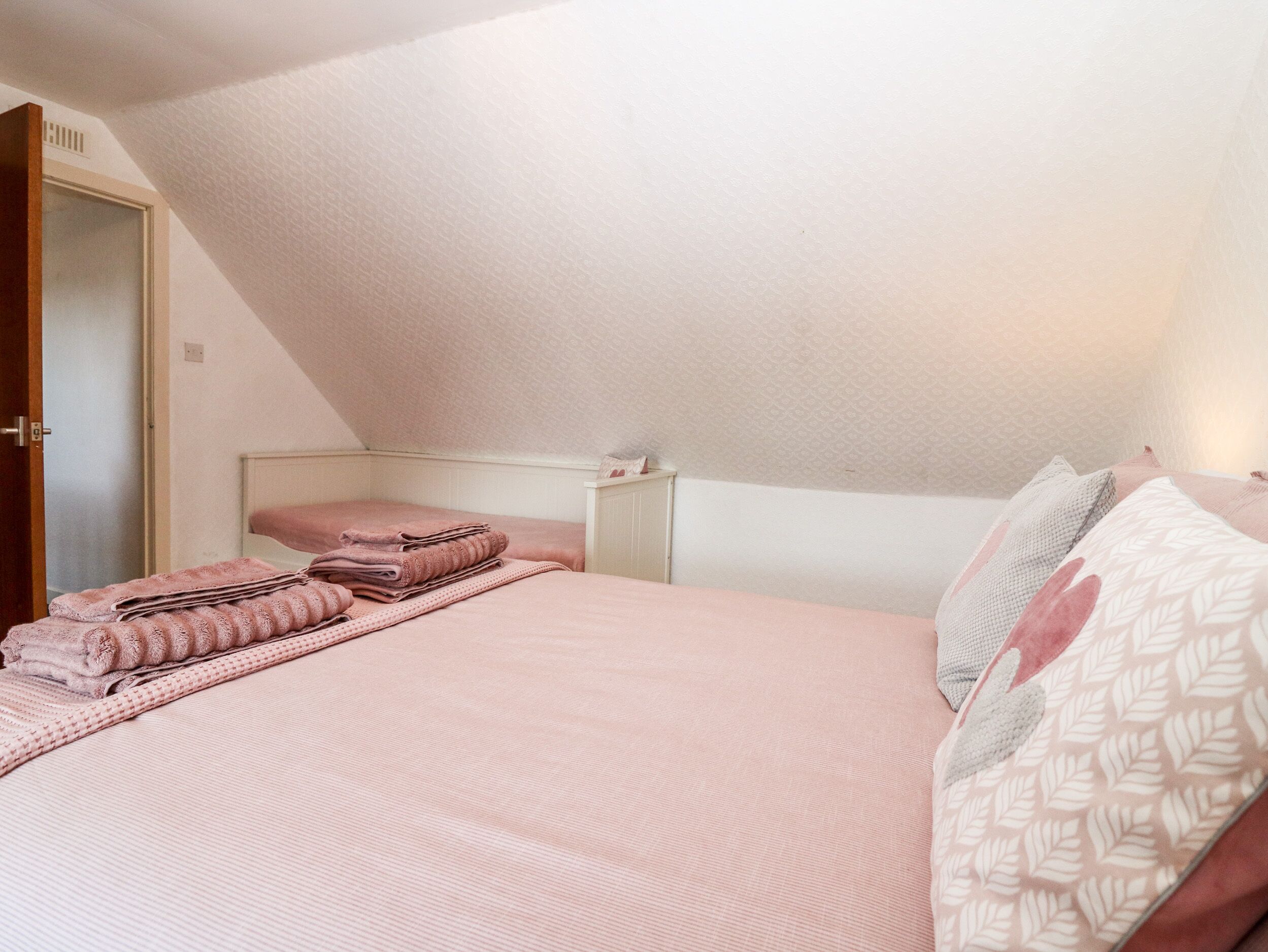 A bedroom with a sloped ceiling, a bed with pink bedding, a nightstand, and a door to the left.