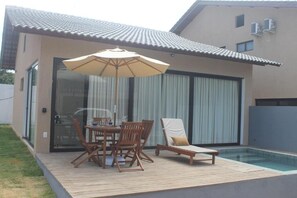 Ein modernes Haus mit Swimmingpool, einer Holzterrasse und einer Glastür, die auf eine Terrasse mit Tisch und Stühlen führt.
