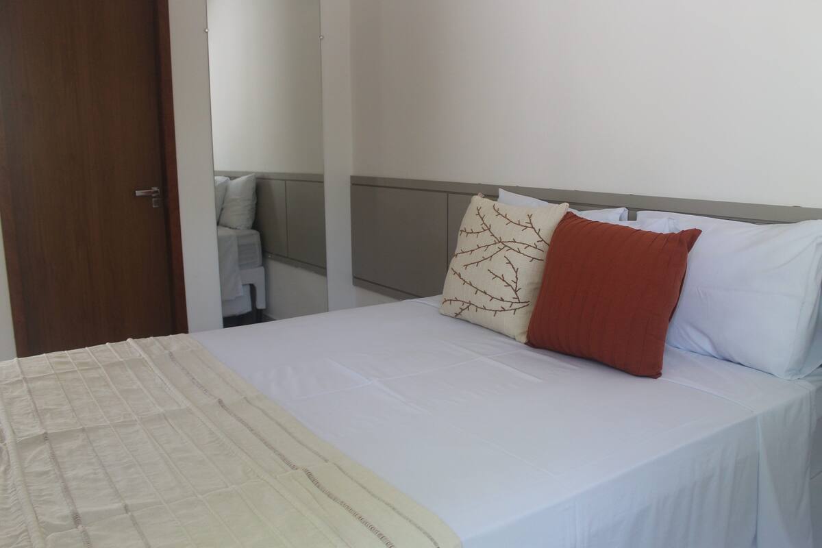 A neatly made bed with white bedding and two pillows, one with a branch pattern and another solid red.