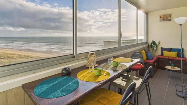 Quarto com uma grande janela com vista para a praia, mesa de jantar com jogos americanos, cadeiras e um sofá com almofadas.