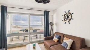 Sala de estar com sofá marrom, mesa de centro branca e vista para a praia através de grandes janelas.