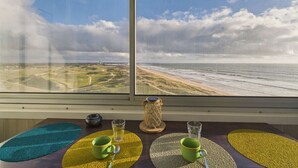 Quarto com uma grande janela com vista para a praia e oceano, uma mesa de madeira com uma caneca verde, um copo de vidro transparente e um objeto decorativo.