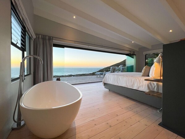 A modern bedroom with a large bathtub, a bed with pillows, and a view of the ocean.