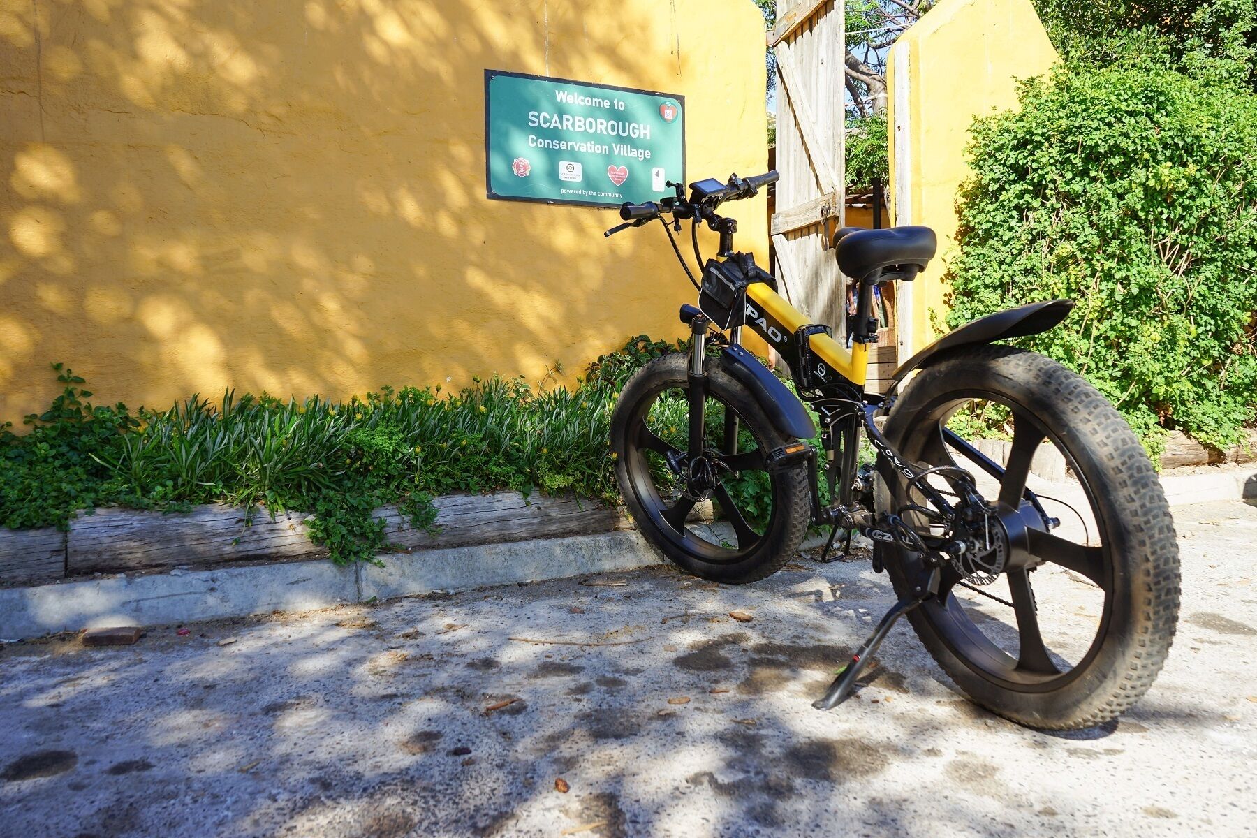 A bicycle parked against a yellow wall with a sign that reads 'Welcome to Scarborough Conservation Village.'