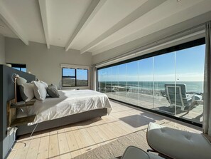 A modern bedroom with a large bed, a nightstand, a lamp, and a view of the ocean through floor-to-ceiling windows.