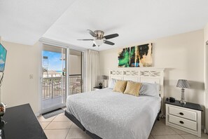 Ein Schlafzimmer mit Bett, Nachttisch, Lampe, Deckenventilator, einem Gemälde und einer Glastür zum Balkon.