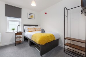 Une chambre à coucher comprenant un lit, une table de chevet, une fenêtre avec une plante, un radiateur et une étagère fixée au mur.