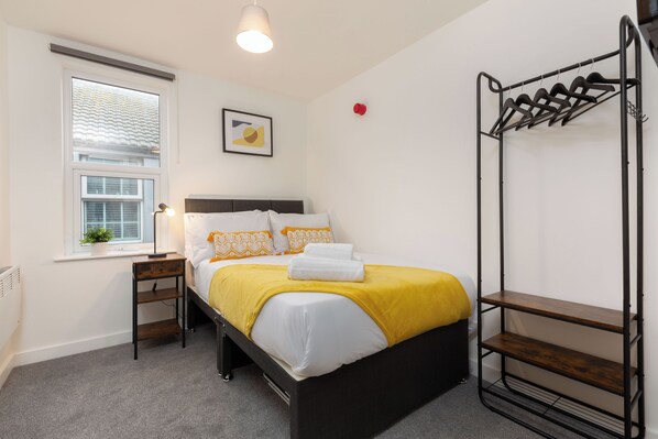 A bedroom with a bed, a bedside table, a window with blinds, a wall-mounted shelf, and a red object on the wall.