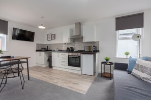 A modern kitchen with white cabinets, a black countertop, and a built-in oven. There is a dining area with a table and chairs, and a gray sofa with cushions.