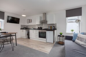 A modern kitchen with white cabinets, a black countertop, and a built-in oven. There is a dining area with a table and chairs, and a gray sofa with cushions.
