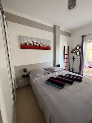 Una cama bien hecha con una colcha a rayas, una mesita de noche con una lámpara, un estante en la pared con objetos decorativos y una ventana con cortinas.