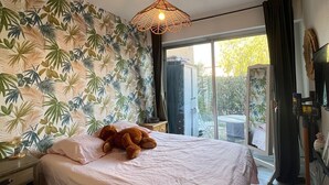 Une chambre à coucher avec un lit, un ours en peluche, un luminaire au plafond, une fenêtre et un mur orné d’un motif de feuilles tropicales.