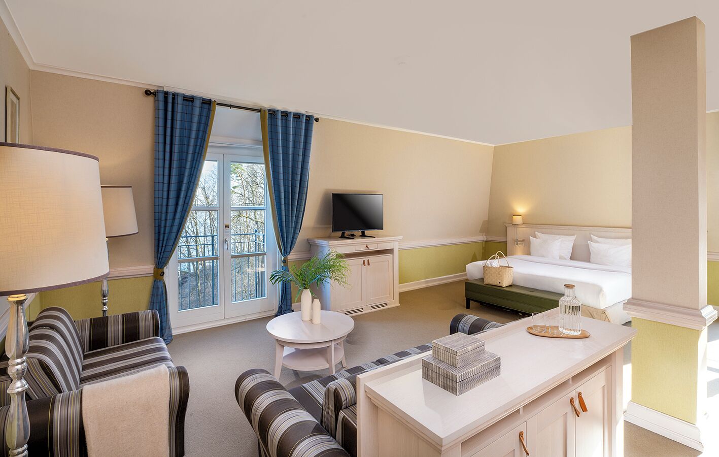 A hotel room with a bed, a TV, a desk with a lamp, a chair, a sofa, a small table with a plant, and a window with curtains.