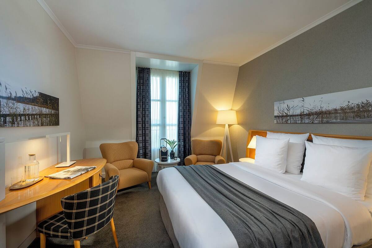 Una habitación de hotel con una cama, dos sillones, un escritorio con lámpara, una mesita con una planta y una ventana con cortinas.
