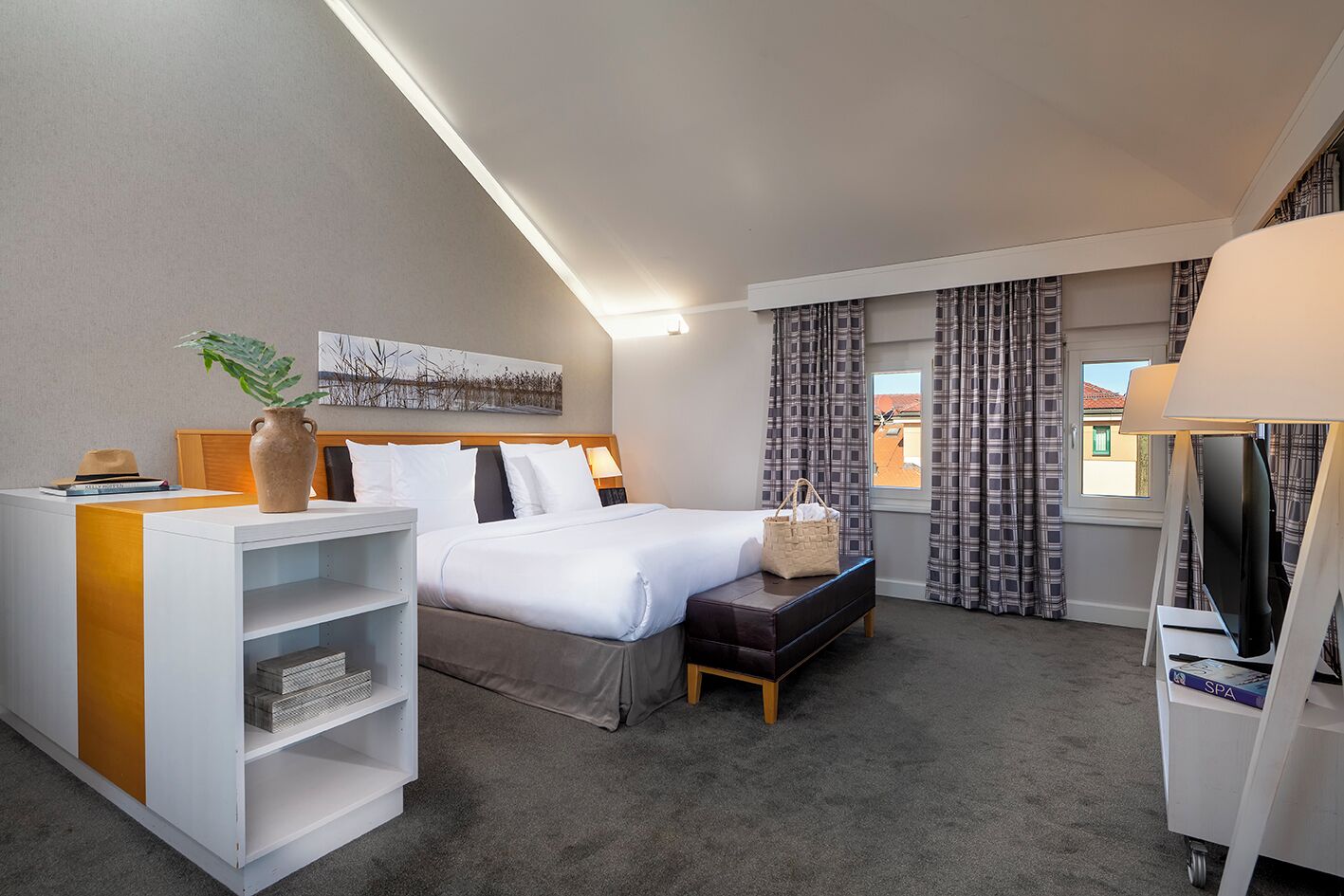 Une chambre d’hôtel avec un lit, un bureau doté d’une lampe, une télévision, une fenêtre avec des rideaux et une plante.