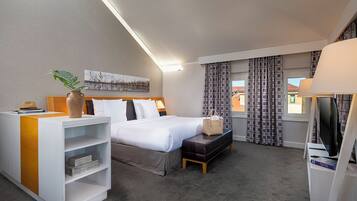 Quarto de hotel com cama, secretária com abajur, televisão, janela com cortinas e uma planta.