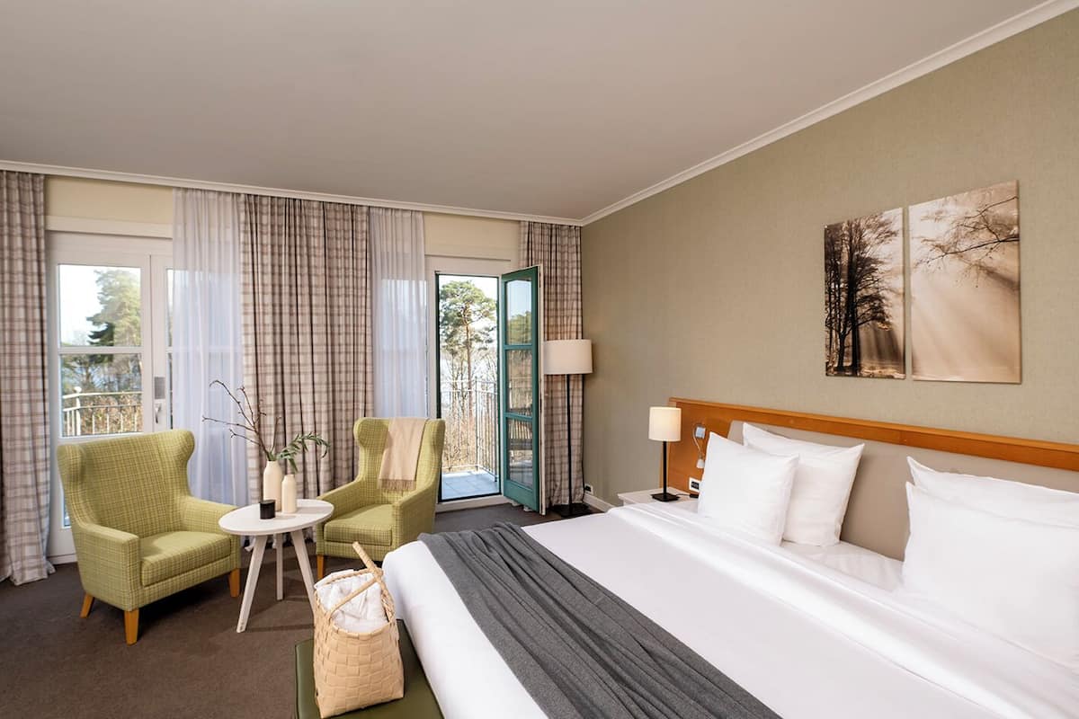 Habitación de hotel con una cama grande, dos sillones, una mesita y vistas al exterior a través de una puerta.
