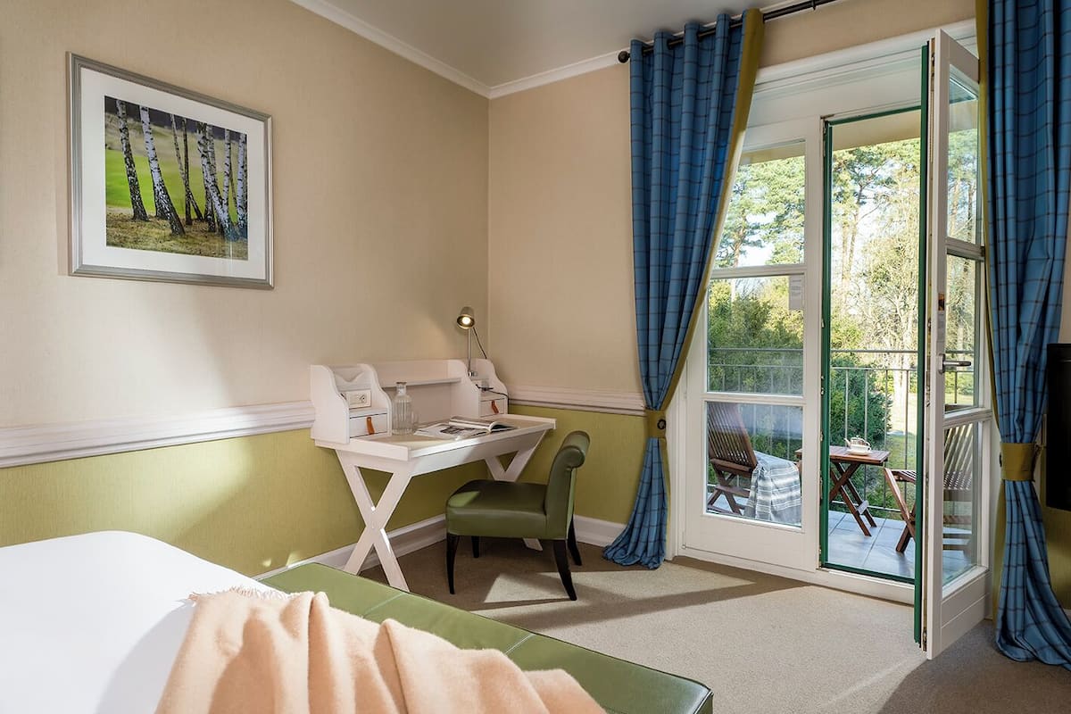 Una habitación con cama, escritorio y silla, ventana con cortinas, y un cuadro en la pared.