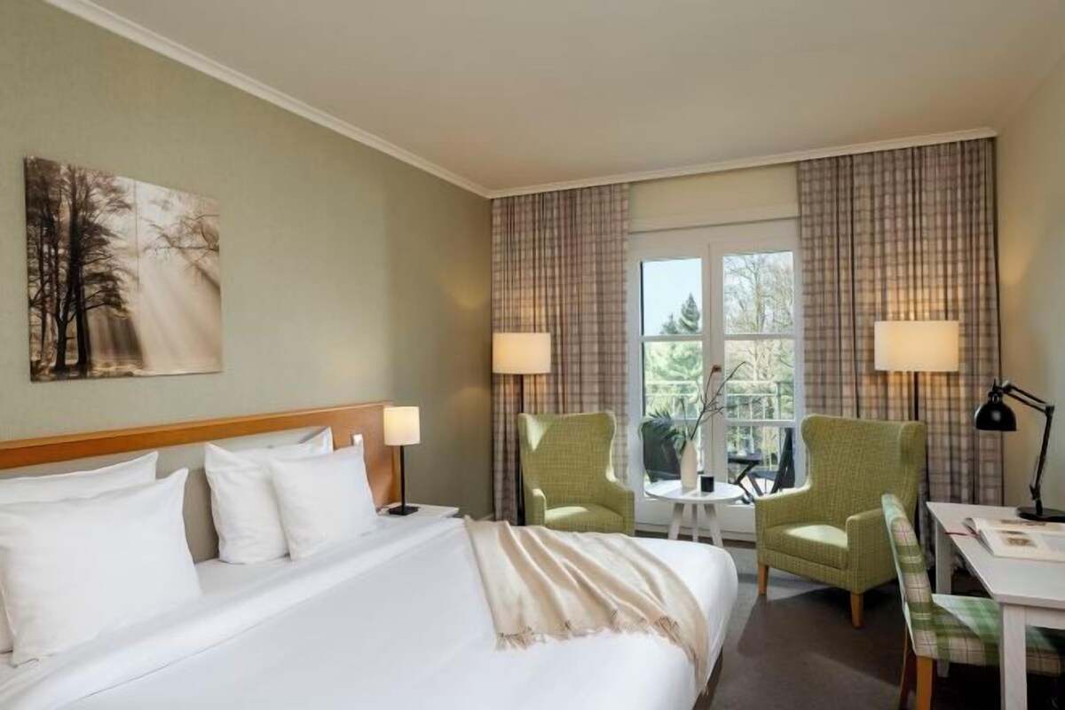 Una habitación de hotel con una cama, dos sillas, una mesita y un ventanal con cortinas.