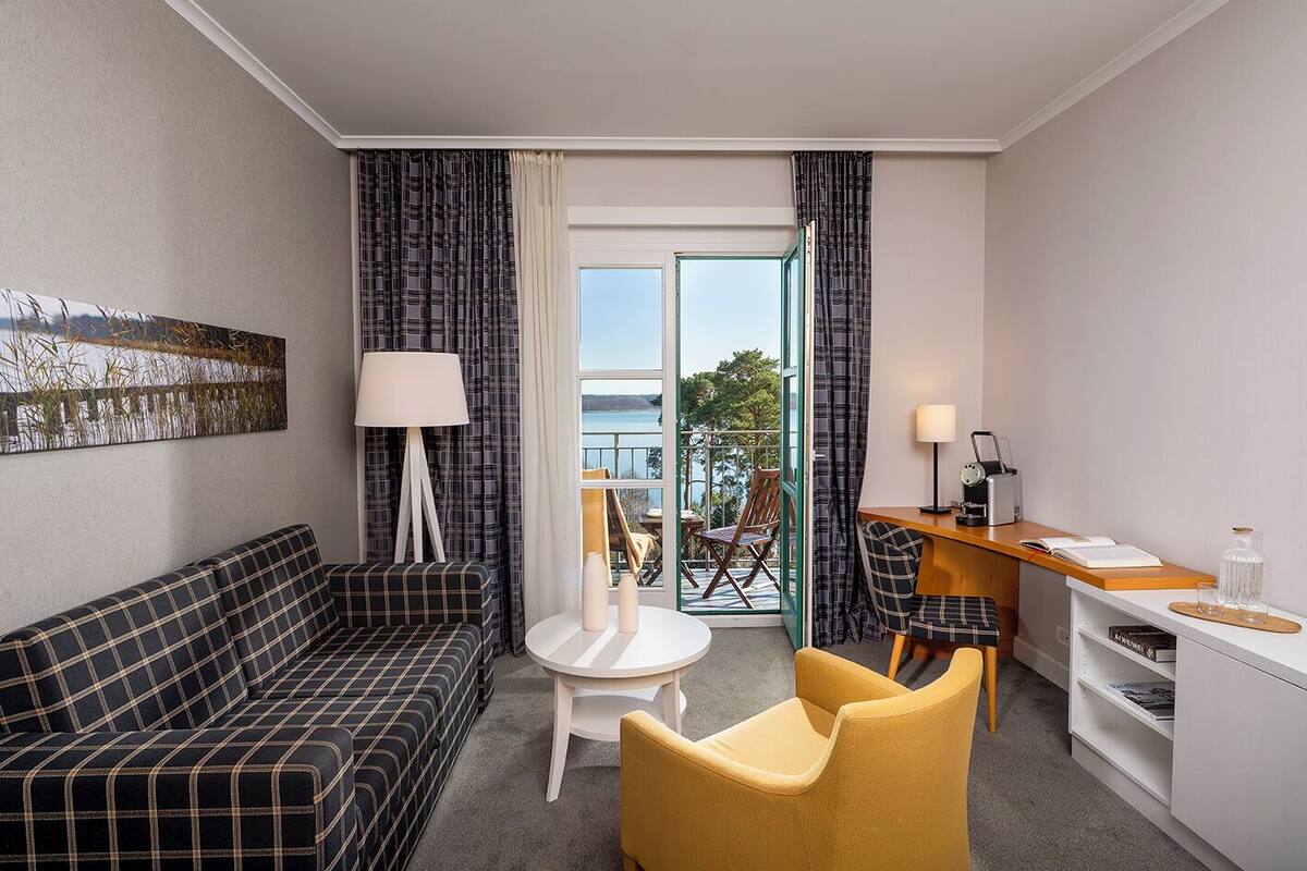 Habitación de hotel con sofá a cuadros, una pequeña mesa redonda, dos sillas, un escritorio con lámpara y vistas al exterior a través de un ventanal.