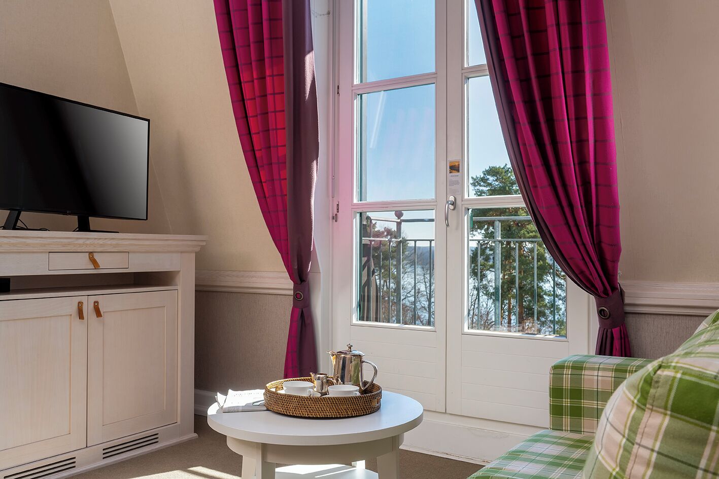 A room with a television on a cabinet, a small round table with a tray, and a view of the outdoors through a window with curtains.