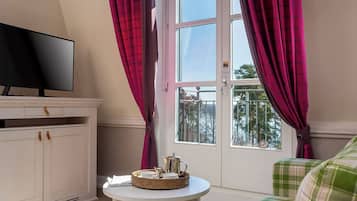 A room with a television on a cabinet, a small round table with a tray, and a view of the outdoors through a window with curtains.
