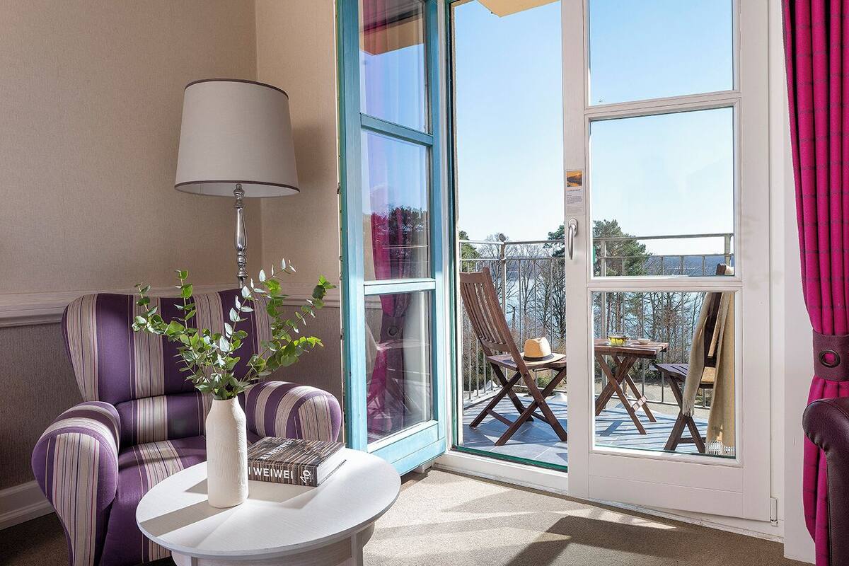 Una habitación con un sillón a rayas, una mesa blanca con un jarrón y libros, una lámpara y un ventanal corredero que da a una zona de estar exterior con una mesa y sillas.