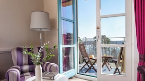 Una habitación con un sillón a rayas, una mesa blanca con un jarrón y libros, una lámpara y un ventanal corredero que da a una zona de estar exterior con una mesa y sillas.